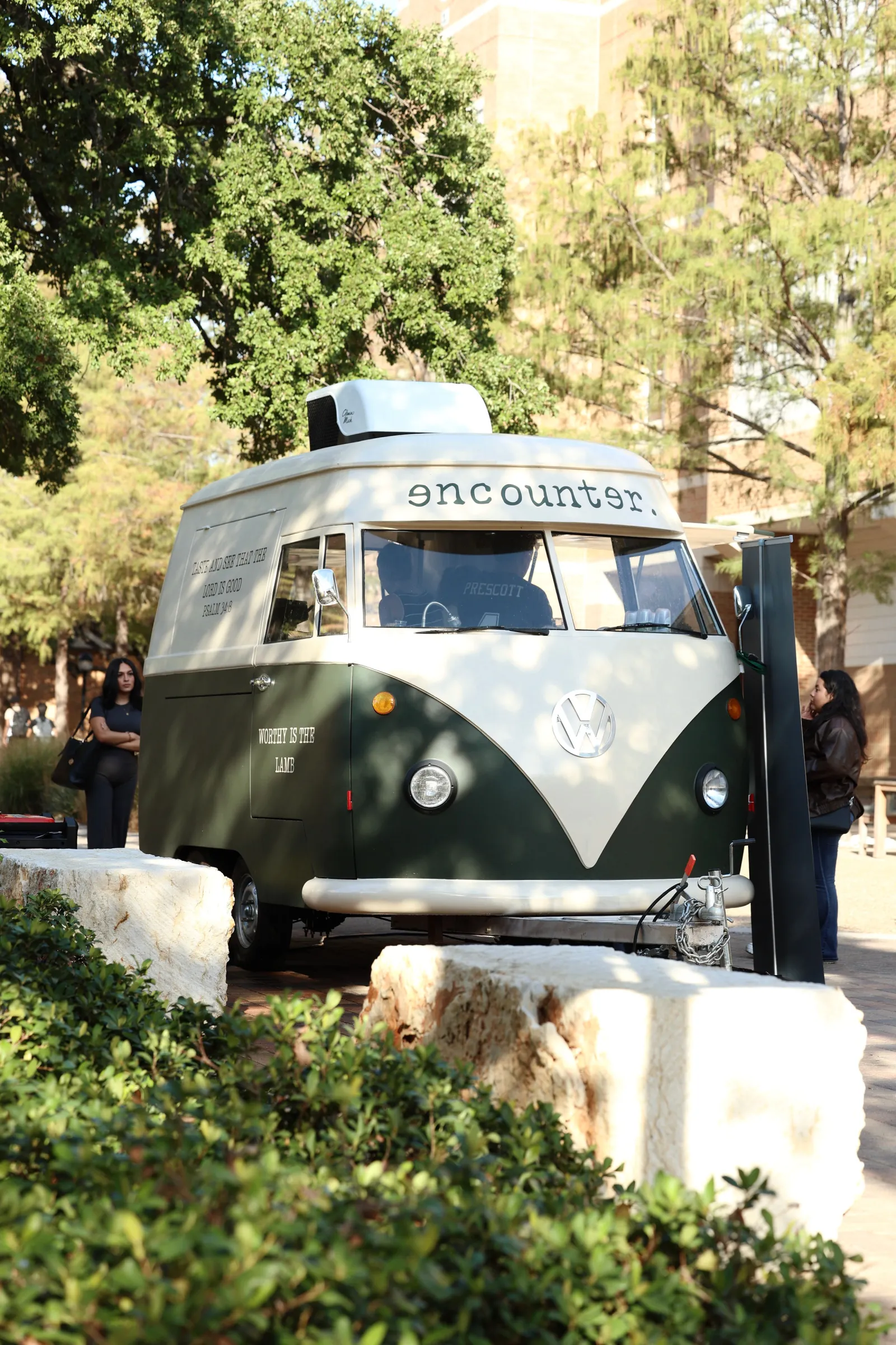 The green and cream Encounter Coffee VW bus coffee trailer parked under the trees at an outdoor North Texas event